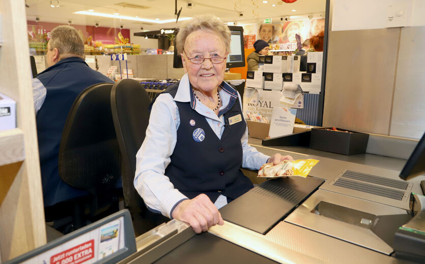 Ehrung für dienstälteste Kassiererin: ältere Frau sitzt an der Kasse und lächelt in die Kamera.