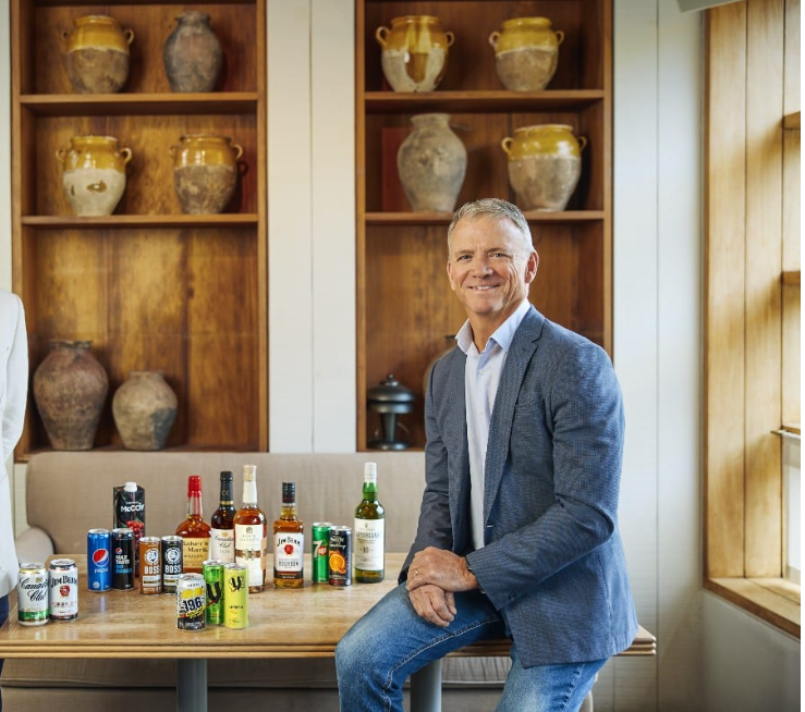 Führungswechsel bei Suntory: Auf dem Foto sieht man rechts im Bild Darren Fullerton. Er sitzt auf einem Tisch, auf dem die Produkte von Sutory stehen. 