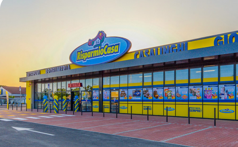 Außenansicht einer Risparmio-Casa-Filiale in Italien mit gelb-blauer Fassade und Schaufensterplakaten bei Sonnenuntergang.