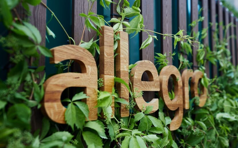 Holzlogo von Allegro an einer Fassade von grünen Kletterpflanzen umrankt.