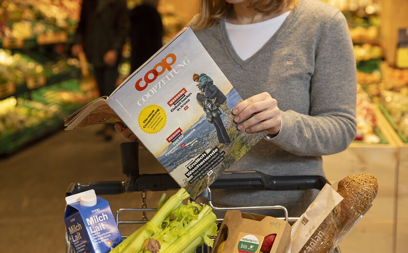 Coop verlegt Start seiner wöchentlichen Aktionen: Eine Frau schaut in das Coop Kundenmagazin. Vor ihr steht ein gefüllter Einkaufswagen.