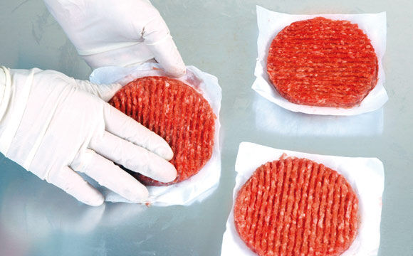 Hand mit Handschuhen legt rohes Burger-Patty aus Hackfleisch auf Papier.