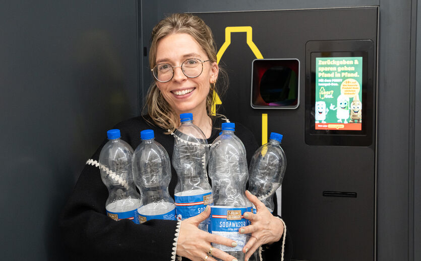 Penny Österreich belohnt Pfandrückgabe mit Rabatt: Eine Frau steht lachend vor einem Pfandautomaten und hält leere Plastikflaschen im Arm.