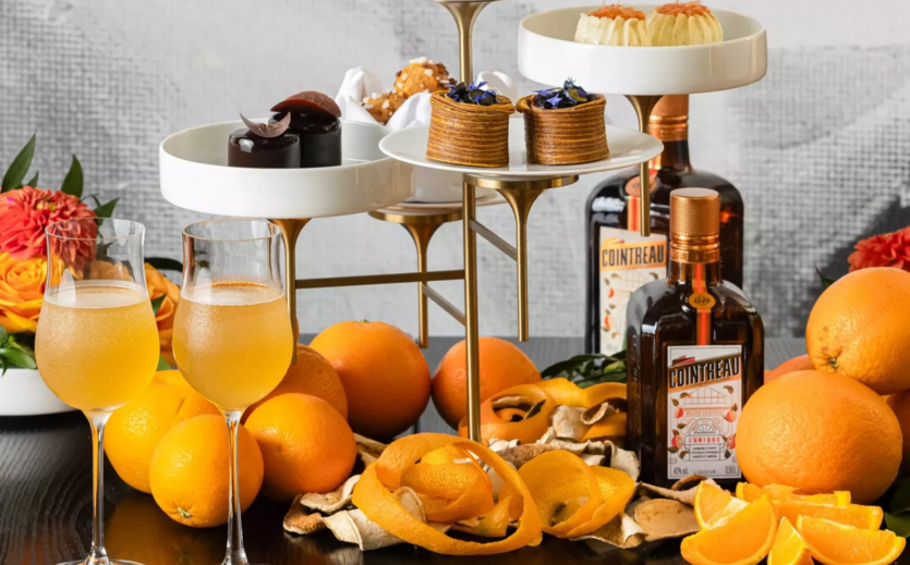 Flaschen Cointreau-Likör, Orangen, Dessertvariationen und zwei Cocktailgläser auf einem gedeckten Tisch.