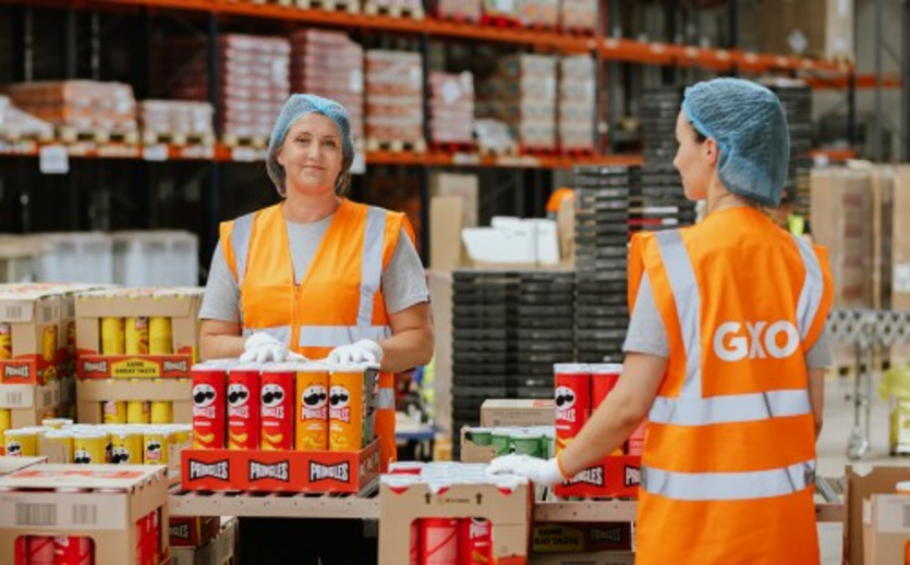 Zwei Mitarbeitende von GXO arbeiten in einem Logistikzentrum zwischen Paletten und Waren.
