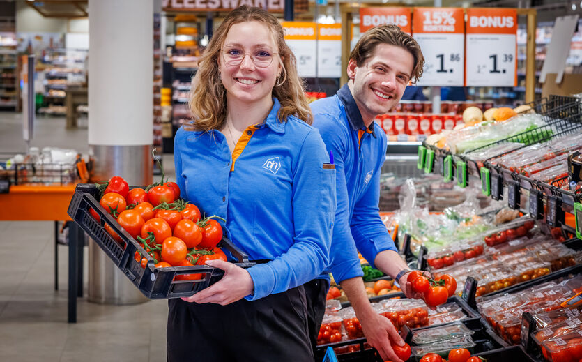 Albert Heijn steigert Bio-Anteil auf 10 Prozent: Eine junge Albert Heijn Mitarbeiterin hält eine Steige mit Tomaten. Ein junger Kollegen steht hinter ihr vor der Gemüseauslage.