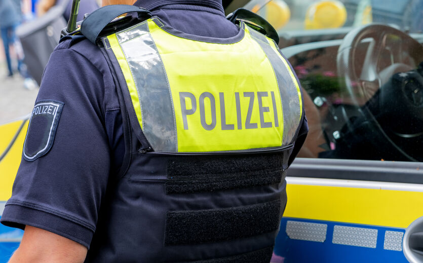 Schüsse auf Spätkauf-Geschäfte: Polizist von hinten in gelter Weste mit Polizei-Schriftzug stet an Streifenwagen.sc