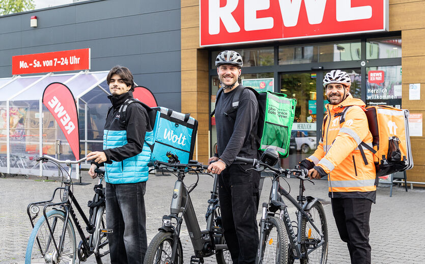 Rewe-Sofort-Lieferung: Drei Fahrer von Wolt, Uber Eats und Lieferando stehen vor einem Rewe-Markt.