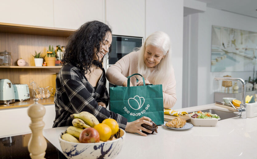 Umfrage zur Esskultur: JUnge Frau und ältere Frau stehen in einer Küche und packen aus einer Too-Good-To-Go-Tüte Lebensmittel aus.
