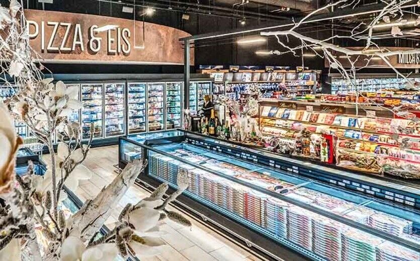 Tiefkühlbereich eines Supermarkts mit Kühltruhen für Pizza und Eis sowie beleuchteten Tiefkühlregalen mit verpackten Lebensmitteln.