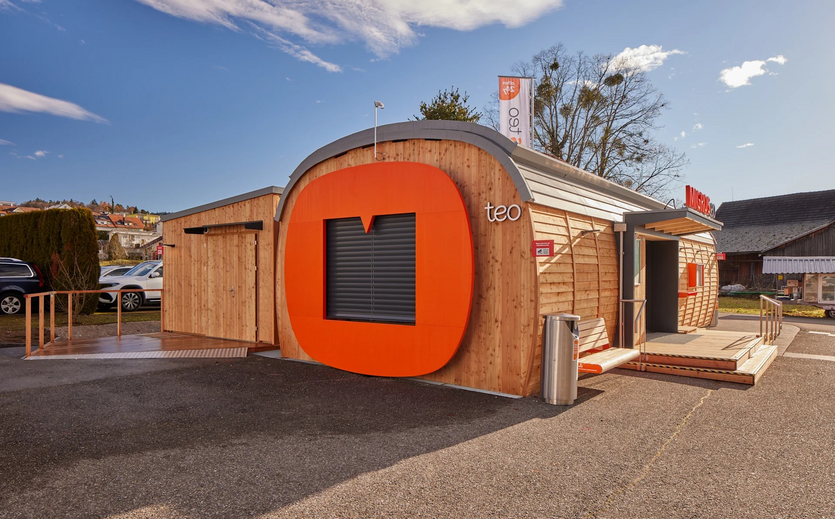 Migros teo Filiale als kleiner Holzbau mit orangefarbenem Logo.
