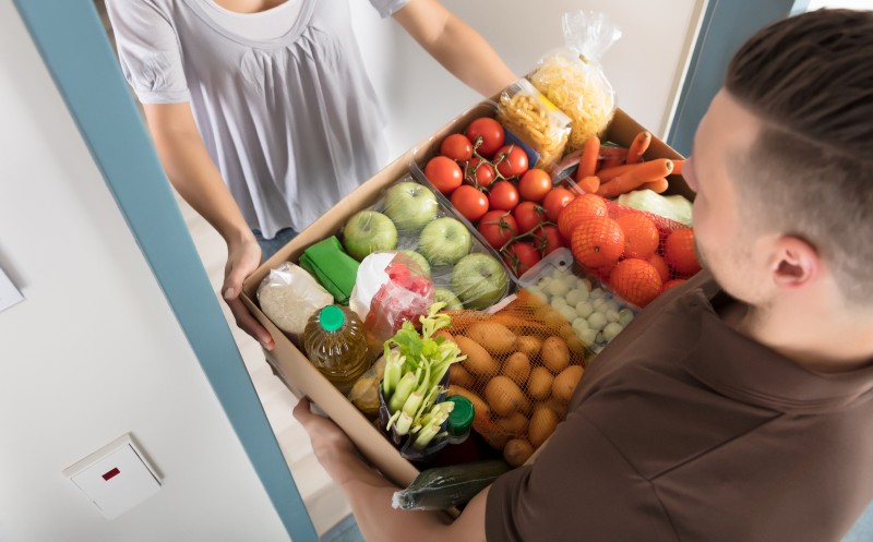 Elevated View Of Smiling Young Woman Accepting Cardboard Box Full Of Groceries From Delivery Man